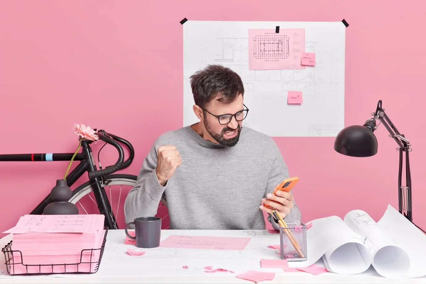 An outraged, angry male office worker glares at his smartphone in annoyance, fist clenched, distracted from work, posing in a coworking space.