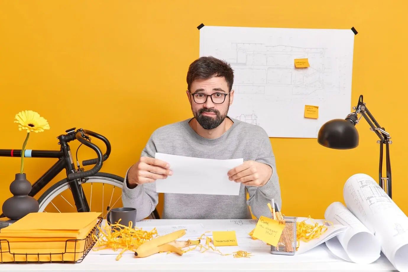 A puzzled, bearded, skilled male illustrator holds paper, having trouble with a future project, posing in a coworking space with a messy desk. A male freelance architect is preparing drawings for a new house development.